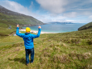 Fototapeta na wymiar Male tourist in blue jacket and yellow hat looking at stunning nature scene with beach, ocean, mountain and cliffs. Warm sunny day. Keem bay, Ireland. Travel and tourism. Irish nature landscape.