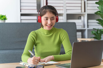 Young asian woman in wireless headphones studying online, using laptop and taking notes while sitting in living room at home.