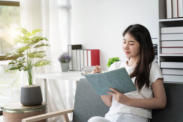 Attractive young Asian woman  sitting on the sofa in the minimal and comfortable living room enjoying reading a book.