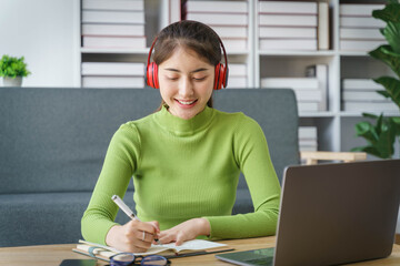 Young asian woman in wireless headphones studying online, using laptop and taking notes while sitting in living room at home.