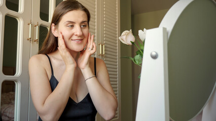 Elegant woman checking her face for wrinkles at mirror in bedroom. Skin care, female health and beauty.