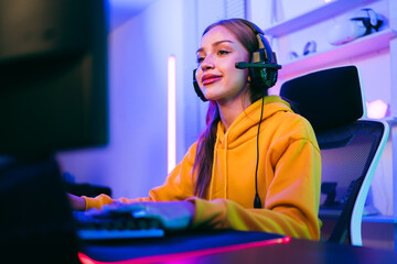 Young Caucasian woman professional gamer wearing yellow hoodie sits on a chair with a gaming table with pc, keyboard, monitor, microphone. Prepare for competition, cast gameplay or record a podcast.