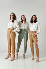 Full-length portrait of three beautiful women in stylish clothes, pants and blouses standing against light studio background. Concept of elegant fashion, female beauty, business, shopping, sales, ad