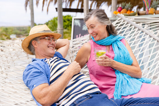 Caucasian senior woman holding drink and lying with husband on hammock at beach