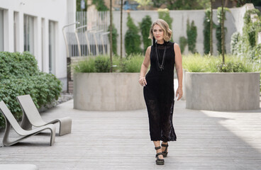 Portrait of Beautiful White Caucasian Adult Woman She is Wearing Black Dress and Bijouterie on Neck. Vilnius Old Town in Background. Flowers and Green Grass in Background.