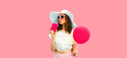 Colorful portrait of beautiful young woman drinking fresh juice with yellow balloon wearing white summer straw hat on blue background