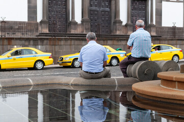 Vista de una céntrica plaza de Funchal, Madeira, con taxis aparcados y dos señores en una fuente.