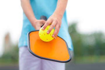 Pickleball paddle and yellow ball close up in children hands, leisure outdoor sport activity.