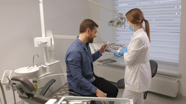 Young attractive man visiting dentist, sitting in dental chair at modern light clinic. Young woman dentist holding x ray image
