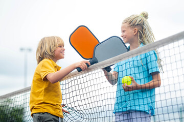 Laughing boy and girl playing pickleball game, hitting pickleball yellow ball with paddle, outdoor sport leisure kids activity.