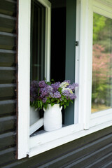A big white ceramic vase with purple and white lilac flowers stands on an old wooden house's window.