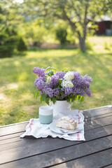 Coffee time with cake and lilac bouquet in a beautiful garden. The background shows a softly blurred tree and lush greenery, suggesting a serene outdoor atmosphere.