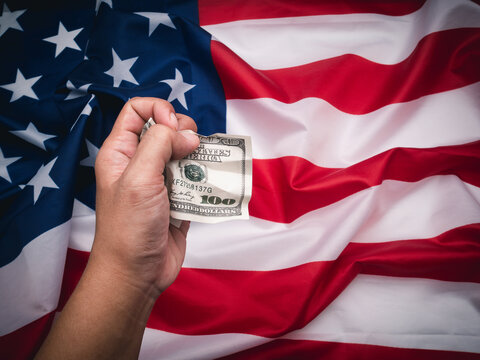 Hand holding one hundred US dollar banknote crumpled over the American flag background.
