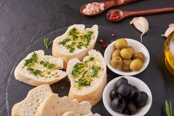 Mediterranean snacks set. Olives, oil, herbs and sliced ciabatta bread on black slate stone board over painted dark blue background, top view. Flat lay.