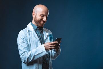 Photo of cheerful doctor with beard smiling and chatting at smart phone with blue background and medical white coat. Medical professional texting sms on cellphone using a social app