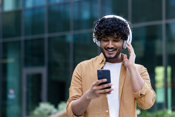 A young male Hispanic student is standing on the street near the campus wearing headphones with a phone in his hands. Listens to music, a podcast, talks on a video call