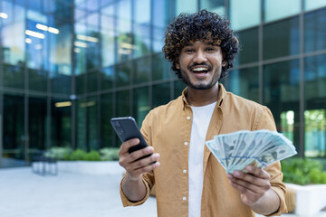 Portrait of a happy young man standing outside an office business center, holding a mobile phone...