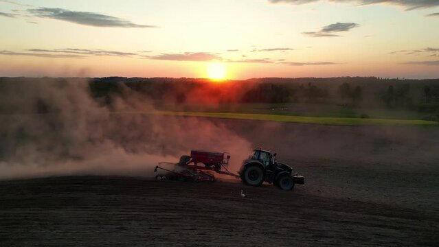 Drone flying over tractor with harrow system plowing ground on cultivated farm field, pillar of dust trails behind with sunset. Tractor plowing the dusty arid soil over agricultural soybean field