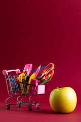 Close up of miniature shopping trolley with school materials, apple and copy space on red background