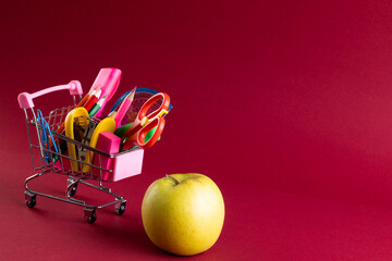 Close up of miniature shopping trolley with school materials, apple and copy space on red background