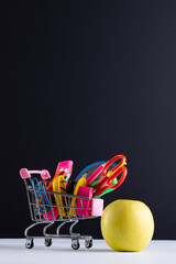 Close up of miniature shopping trolley school materials, apple and copy space on black background