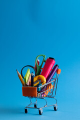 Close up of miniature shopping trolley with school materials and copy space on blue background
