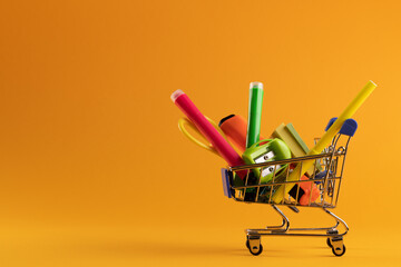 Close up of miniature shopping trolley with school materials and copy space on orange background