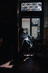 Fototapeta na wymiar Young girl in white dress drinking coffee in a coffee shop near the window in the sun. Silhouette.
