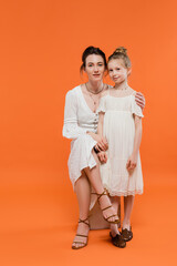 motherly love, brunette young woman sitting on chair and hugging preteen daughter on orange background, white sun dresses, summer fashion, togetherness, love, female bonding, modern parenting