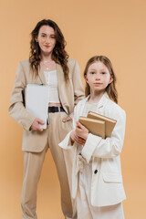 Fototapeta premium trendy mother and daughter in suits, child holding books near blurred woman with notebooks on beige background, fashionable outfits, formal attire, corporate mom, education concept