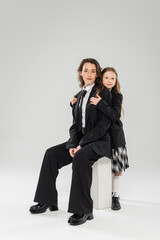 happy girl hugging mother, businesswoman sitting on concrete stool near schoolgirl in uniform on grey background in studio, formal attire, parent-child, modern parenting, fashion shoot