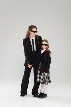 modern parenting, happy businesswoman in suit and sunglasses hugging daughter in school uniform with plaid skirt and standing together on grey background, mother and child