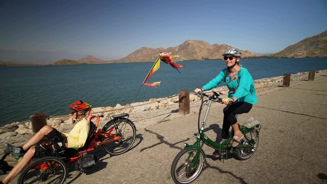 Elderly mother and mature daughter riding e-bikes beside a lake. Mother is on a recumbent electric bike.