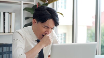 Exhausted young asian worker yawning at workplace. Business man using laptop for working feeling...
