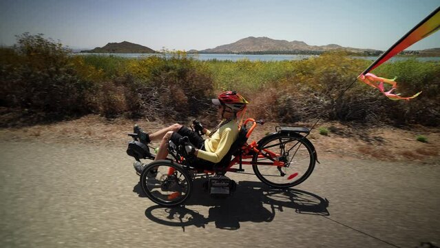 Side view of elderly senior woman riding recumbent e-bike electric tricycle bicycle on a path on a sunny day beside a lake with mountains in the distance.