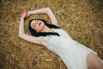 A young girl in a white dress lies on the straw with her eyes closed. Rest in the countryside. © Oleg Kozlovskiy
