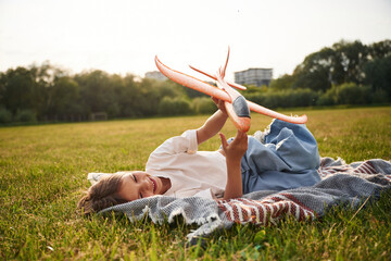 Laying down on the ground. Happy little girl is playing with toy plane outdoors