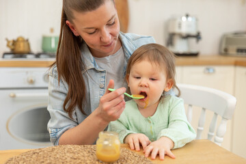 Happy family at home. Mother feeding her baby girl from spoon in kitchen. Little toddler child with messy funny face eats healthy food at home. Young woman mom giving food to kid daughter