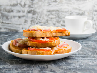 Delicious breakfast, hot sandwiches with tomato, cheese and sausage on a white plate and a cup of coffee on a gray background