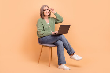 Full length photo of retired woman wear khaki cardigan sit on chair touching glasses hold laptop isolated on beige color background