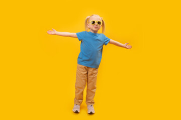 Fashionable little blonde girl in yellow pants and blue T-shirt with stylish sunglasses. Child raised hands up on yellow background and looking at camera.