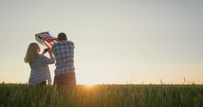Celebrating Independence Day - a man is waving the US flag, a woman nearby is taking pictures with a smartphone. Standing against the backdrop of a field of wheat where the sun sets