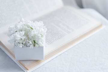 Close-up of a bouquet of white flowers in a white gift box on an open book on a white textile background. Slow life concept. Greeting card mockup	 - obrazy, fototapety, plakaty