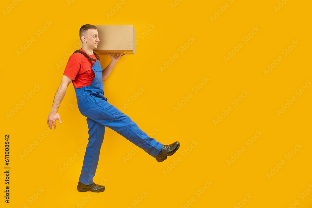 Wall mural Caucasian man in blue uniform carries box on his shoulder. Courier with parcel takes step isolated on yellow studio background. Copy space, mock up.