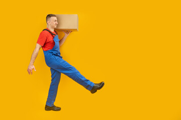 Caucasian man in blue uniform carries box on his shoulder. Courier with parcel takes step isolated on yellow studio background. Copy space, mock up.