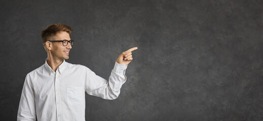 Handsome man advertises or recommends something standing on gray banner background. Young intelligent smiling man points his finger at copy space. Advertising concept. Commercial banner.