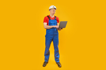 Fototapeta na wymiar Man engineer or worker standing with laptop on yellow background, full-length portrait. Architector in blue overalls, Copy space.