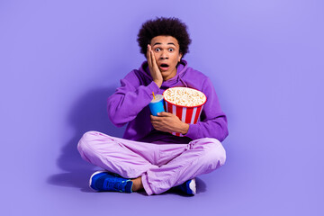 Full length photo of impressed speechless guy wear purple clothes sit hold popcorn drink arm on cheek isolated on violet color background