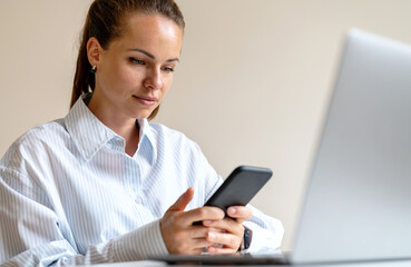 Modern young businesswoman working at office, online messaging using her mobile phone and laptop on the desk.