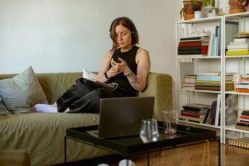 Woman freelancer sitting at her sofa and using smartphone and laptop while working from home office - obrazy, fototapety, plakaty